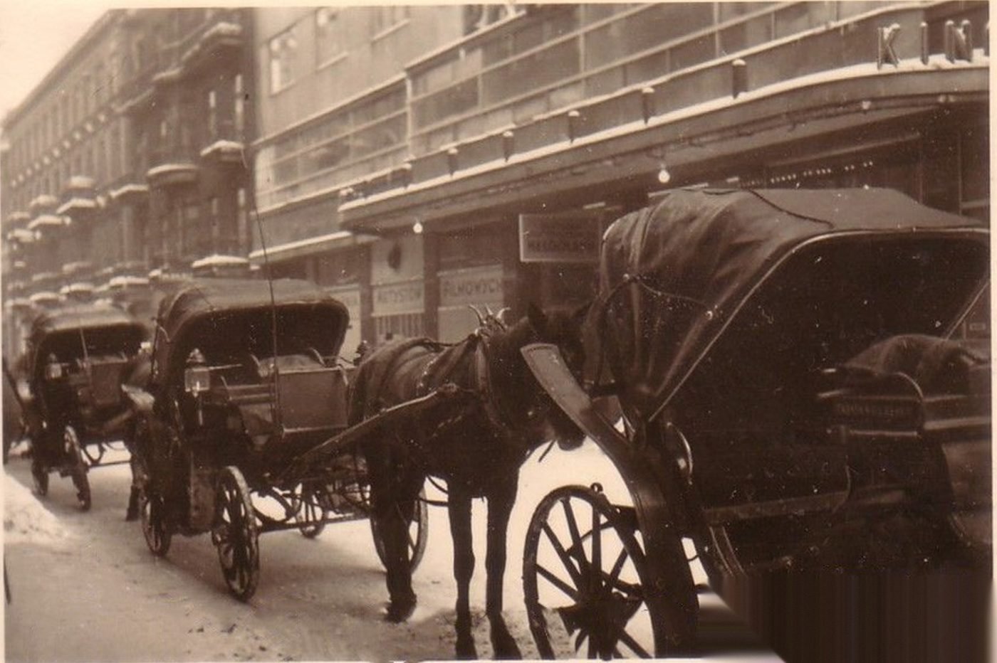 Cabs under the snow-covered marquee of the cinema — winter in occupied Warsaw. The neon signs extinguished; on the window of the premises the inscription „…artystów filmowych" (…of film actors). Photograph after the fotopolska.eu service.