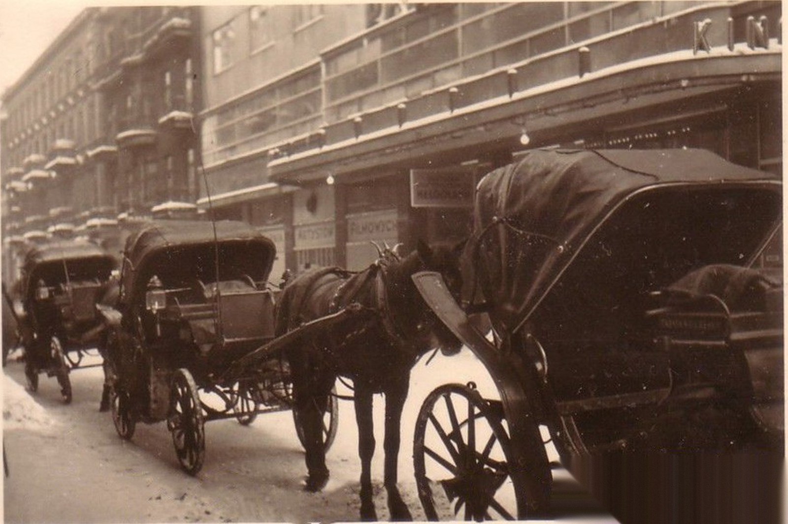 Horse-cabs under the snow-covered marquee of the cinema – winter in occupied Warsaw. The neon signs extinguished; on the shop window the inscription „…film artists". Photograph after the fotopolska.eu service.